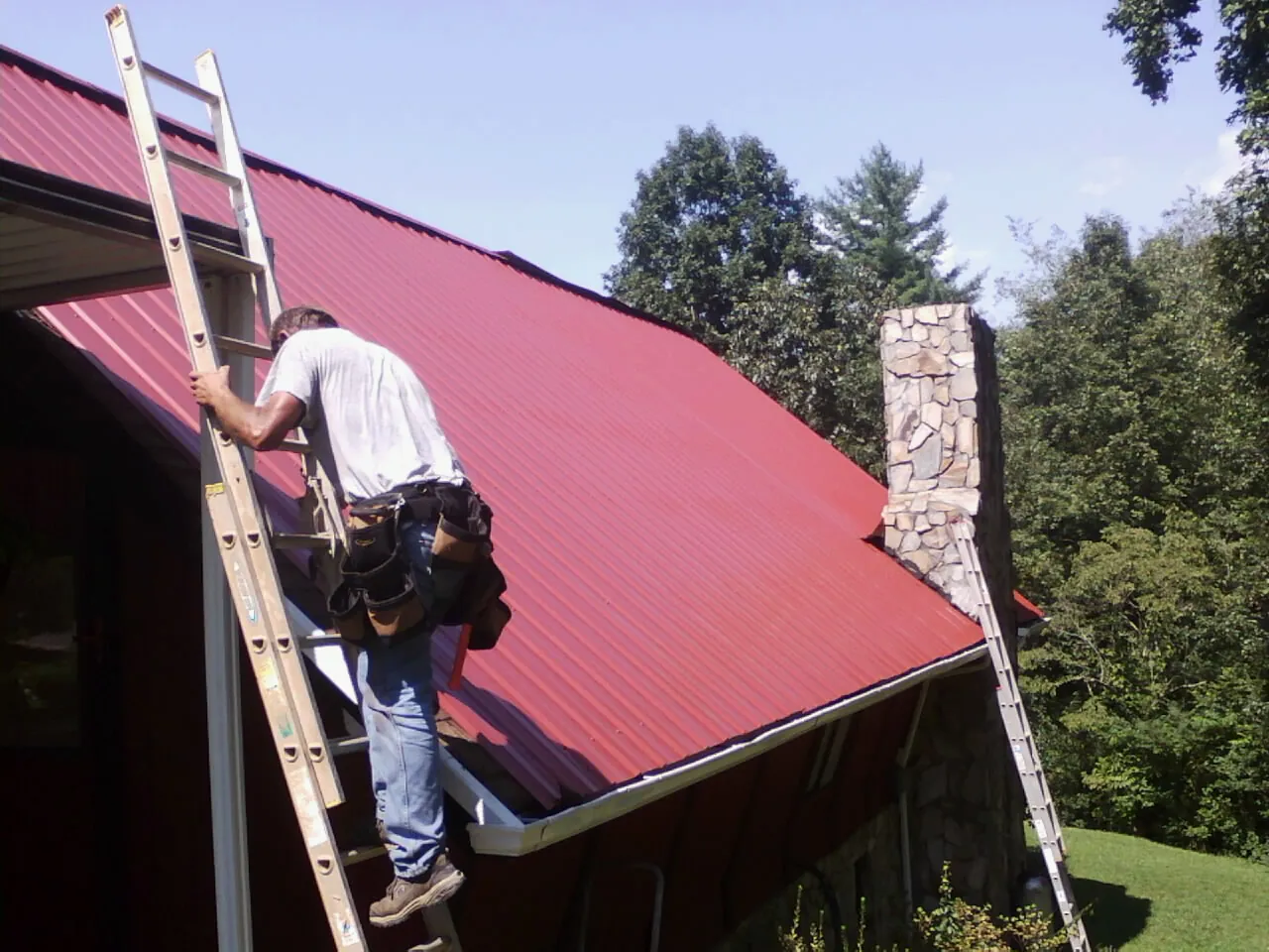 Roofer on a ladder beside a red metal roof and stone chimney — Soffit & Fascia Repair by Roofing Brothers in Jensen Beach